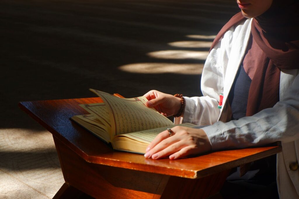 A woman reads the Quran at a wooden stand inside a mosque, bathed in sunlight.